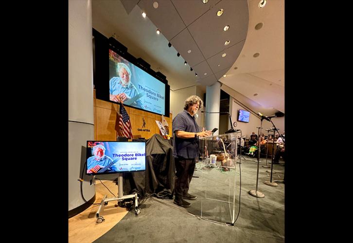 A wide shot of Navarro behind a podium. Navarro is in side view and looks out into the crowd. He wears a dark shirt, black pants and jeans. To the far left of the photo and in the background on a projector is an image of Theodore Bikel and the text, “Theodore Bikel Square” in blue with various logos underneath.