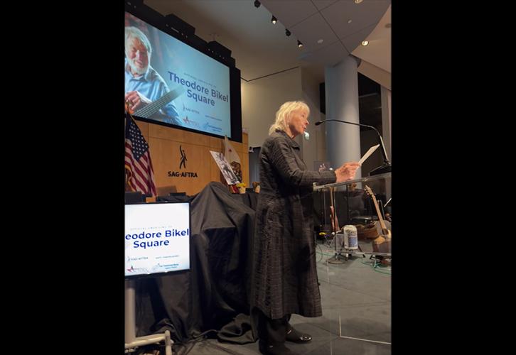 A profile shot of O’Hara behind a podium. O’Hara, looking out into the crowd, wears a long black coat and black slacks and shoes. She speaks into a microphone and holds a sheet of paper. On the projector is an image of Theodore Bikel and the text, “Theodore Bikel Square” in blue with various logos underneath.