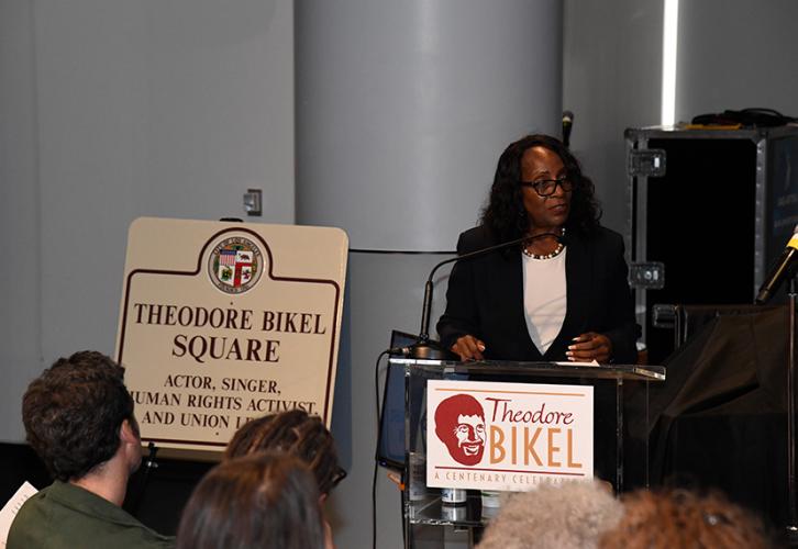 Wheeler stands behind the podium in an auditorium-like room. She wears a black blazer and a white shirt. The podium bears a sign with Bikel’s face and name in stylized lettering. To her right, propped up on an easel, is a large sign of the Theodore Bikel Square.