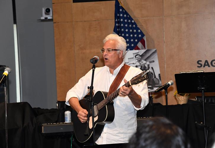 Taubman mid-performance in an auditorium-like room. He wears a white shirt and a guitar strap is over his shoulder. The guitar itself is black with silver detail. Some crowd members are visible in the foreground; behind him is a black-and-white photo of Theodore Bikel and an American flag. There is also a wood-paneled wall; the SAG-AFTRA logo is partially visible.