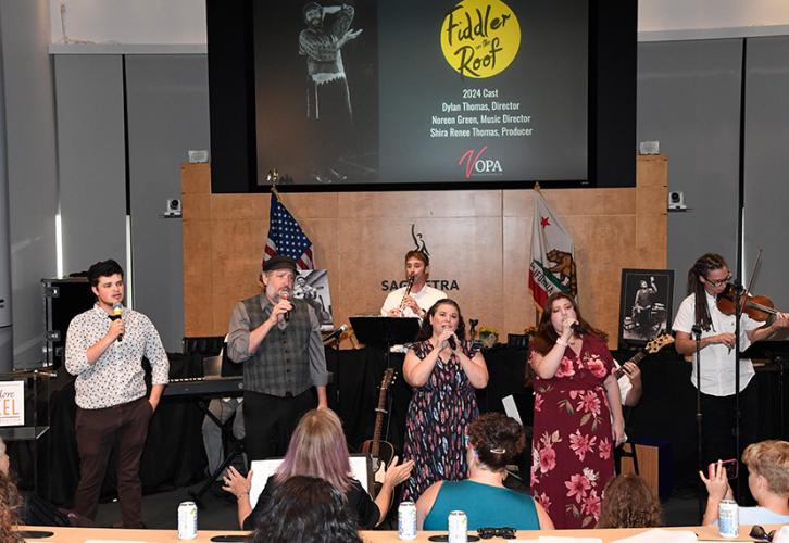 A wide shot of a group of singers, musicians and some of the crowd from the back of an auditorium-like room. The singers, two men and two women, sing into hand-held microphones. Behind them is a clarinet player and a fiddler. Behind them on a projector is a photo of Bikel as Tevya from ‘Fiddler on the Roof,’ artwork from the show and unintelligible text.