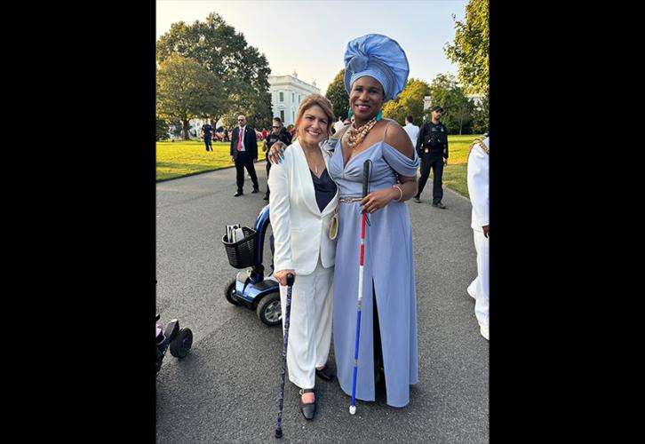 Cyrille con Lachi, una mujer alta con un vestido azul pálido y un peinado a juego. Están en un camino de grava sobre el césped; a lo lejos se ven otros asistentes.