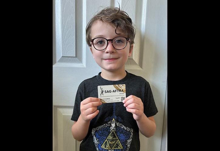 Una persona sostiene su tarjeta de miembro de SAG-AFTRA. Lleva pantalones cortos cargo, una camiseta negra con el símbolo de “Legend of Zelda” dentro de un escudo y gafas. Está de pie dentro de una habitación; detrás de él hay una puerta blanca. La tarjeta es blanca con el logotipo de SAG-AFTRA en negro y pinceladas curvas doradas en las esquinas superior derecha e inferior izquierda.