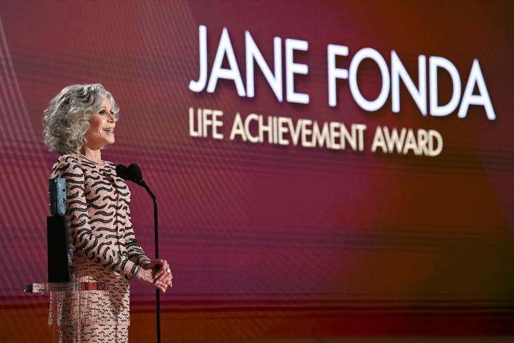 Jane Fonda stands on stage. She speaks into a microphone. Projected on the screen behind her is "Jane Fonda Life Achievement Award" in all-cap. "Jane Fonda" is in white; "Life Achievement Award" is in yellow.