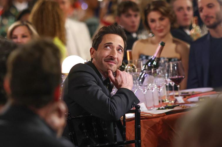 Adrian Brody is seated at a table. He is smiling and looks up. 