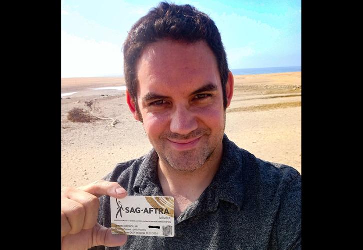 Una persona sostiene en la mano su tarjeta de membresía de SAG-AFTRA. Se encuentra de pie en una zona desértica. Lleva una camiseta de color carbón. La tarjeta es blanca con el logotipo de SAG-AFTRA en negro y pinceladas curvas doradas en las esquinas superior derecha e inferior izquierda.