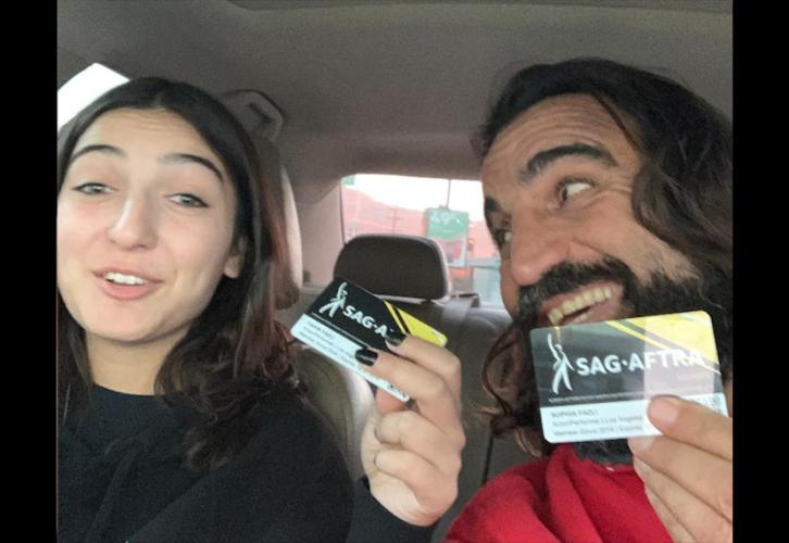 Dos personas sostienen sus tarjetas de membresía de SAG-AFTRA. Están dentro de un automóvil. La persona de la derecha mira a la persona de la izquierda. Sus tarjetas son negras con un triángulo amarillo y una línea amarilla más delgada en la esquina superior derecha y un borde blanco en la parte inferior; el logotipo de SAG-AFTRA se muestra prominentemente en blanco.
