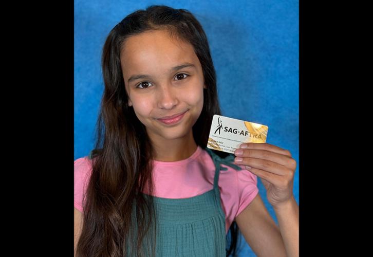 Una persona sostiene su tarjeta de miembro de SAG-AFTRA. Lleva un vestido verde azulado sobre una camiseta rosa. La tarjeta es blanca con el logotipo de SAG-AFTRA en pinceladas curvas negras y amarillas en las esquinas superior derecha e inferior izquierda. El fondo detrás de ellas es azul.
