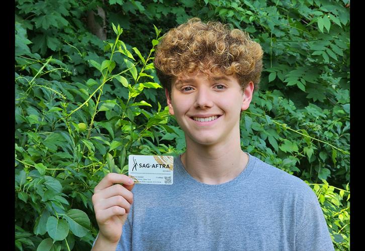 Una persona sostiene su tarjeta de miembro de SAG-AFTRA. Está afuera, entre plantas verdes. Lleva una camiseta gris claro y su cabello es rizado. La tarjeta es blanca con el logotipo de SAG-AFTRA en negro y pinceladas curvas doradas en las esquinas superior derecha e inferior izquierda.
