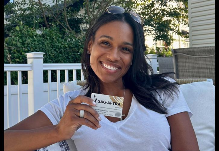 Una persona sostiene su tarjeta de miembro de SAG-AFTRA. Está sentada en un patio al aire libre. Lleva una camiseta blanca, un anillo en el dedo anular derecho y gafas de sol en la cabeza. La tarjeta es blanca con el logotipo de SAG-AFTRA en negro y pinceladas curvas doradas en las esquinas superior derecha e inferior izquierda.