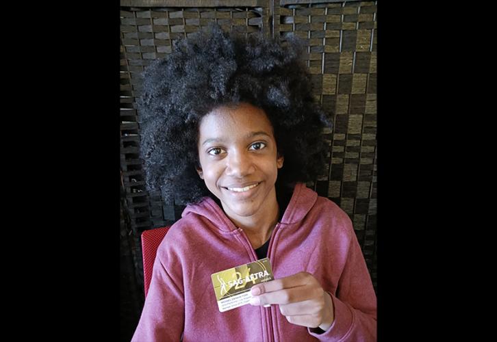 Una persona muestra su tarjeta de membresía de SAG-AFTRA. Lleva una sudadera con capucha roja descolorida y un afro prominente. La tarjeta es amarilla y dorada con un borde blanco en la parte inferior y muestra el logotipo de SAG-AFTRA y el número “24” en amarillo.