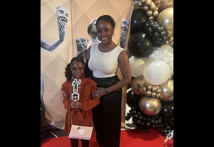 Two individuals, one older and one younger, are on the red carpet. The older individual wears black and white; the younger individual is in a brown dress and holds a party prop, a paper cutout of the SAG Awards’ statuette. Around them are balloons and a banner with the statuette.