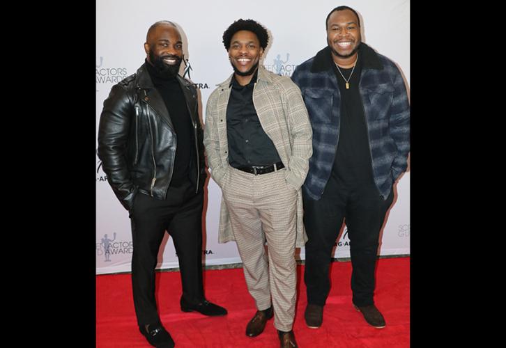 Three individuals are on the red carpet. The one on the far left is in a black leather jacket; the one in the center wears a gray plaid suit; the one on the far right wears a blue flannel jacket with trim. Each has their hands in their pockets. Behind them is the step-and-repeat backdrop.