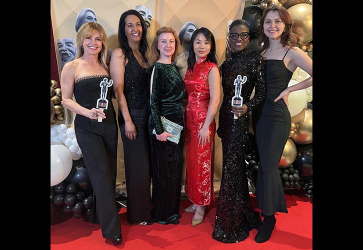 A group of six women in elegant attire pose in front of SAG Awards banners. Two are holding paper cutouts of the SAG Awards statuette. There are also two balloon displays on either side of the group.