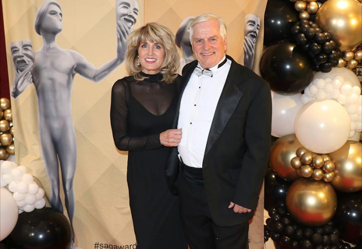 Two individuals are on the red carpet. They are dressed in formalwear, the first in a floor-length black dress and the second in a black tuxedo. Behind them is a SAG Awards banner that is framed with balloons as decoration.