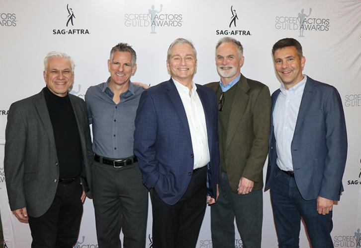 Five men wearing dress shirts and blazers in various shades of blue, gray and green stand shoulder to shoulder for a photo. Behind them is a white step-and-repeat backdrop.