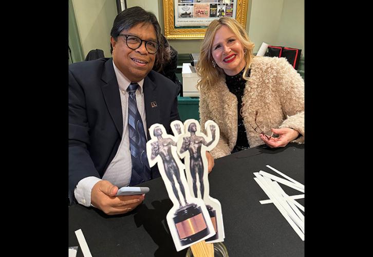 Two people are seated at a small table with a black tablecloth. A jar of SAG Awards props, paper cutouts of the award statuette, sits on the table.