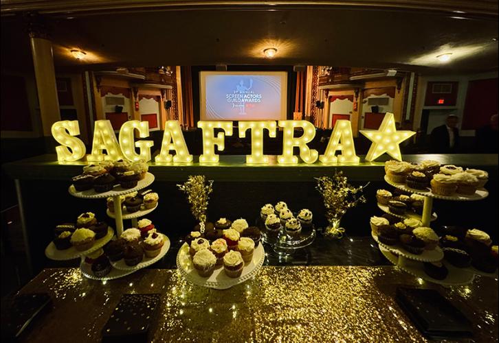 A table with a gold glitter run features cupcake stands with a variety of cupcakes. Some of the stands are multi-tiered. Behind it is a sign that spells “SAG-AFTRA” with a star, and in the background, a projection screen bearing the SAG Awards title card.