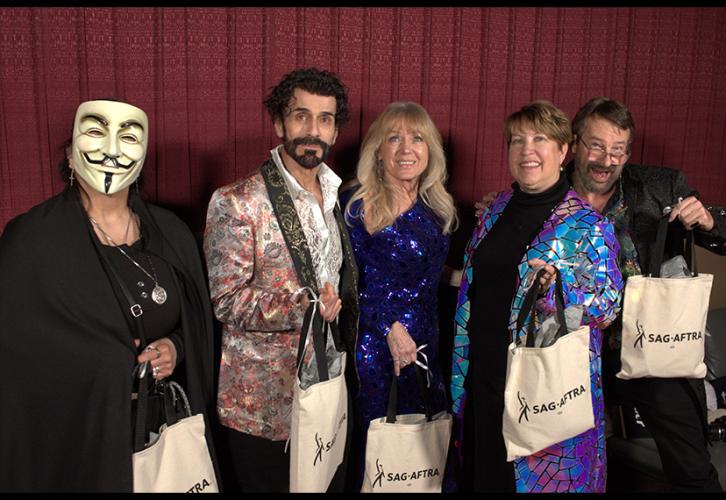 A small group of diverse people dressed in flashy attire holding canvas bags with the SAG-AFTRA logo in front of a movie theater’s red fabric wall. The person on the far left wears a Guy Fawkes mask.