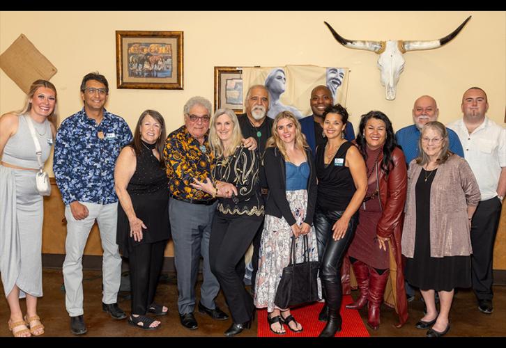 A diverse group of 13 people dressed nicely stand together and smile inside a restaurant room with Southwestern-themed art on the wall behind them. There is also a banner featuring the SAG Awards statuette.
