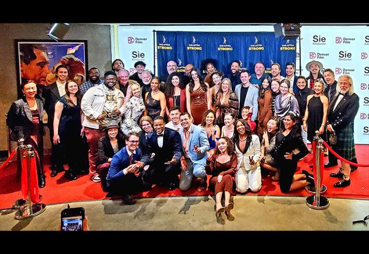 A large group photo inside a building lobby. Many in the foreground are kneeling, while those in the back are standing. Visible is a backdrop with a pattern of the SAG-AFTRA logo and “strong” in all-cap, yellow lettering.