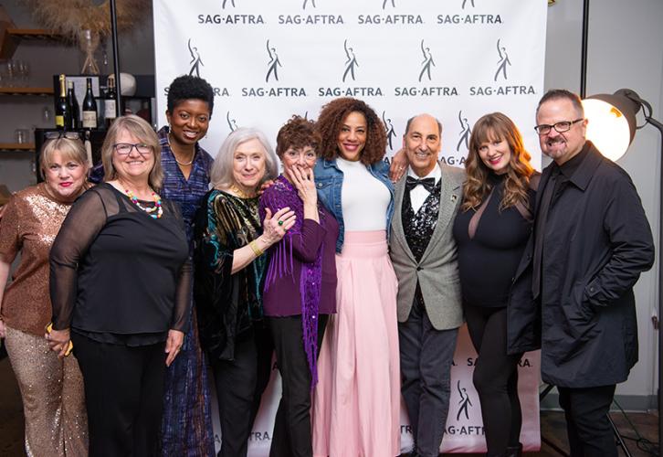 A group photo of nine people inside a bar or restaurant-like setting. Behind them is a white step-and-repeat backdrop with the SAG-AFTRA logo. 