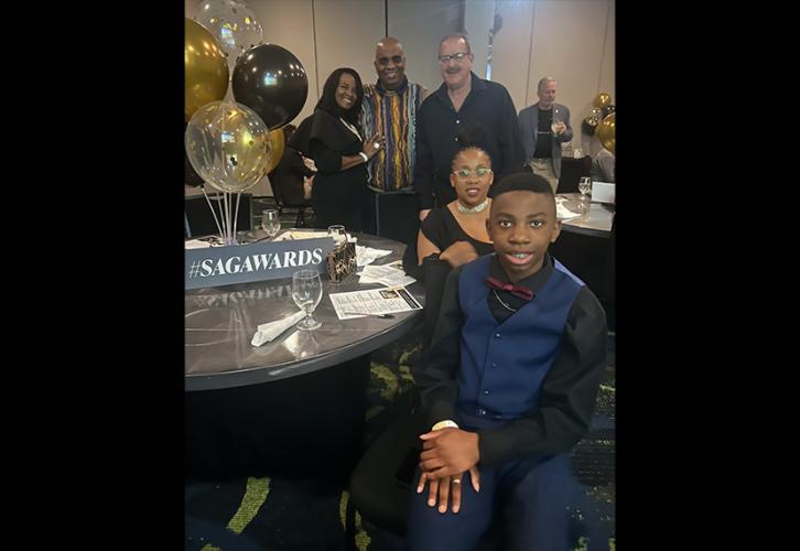 Five individuals are around a table. Three people in the back are standing; two people are seated. On the table is a centerpiece with balloons and a dark gray placard with “#SAGAwards” in all-cap, white lettering.