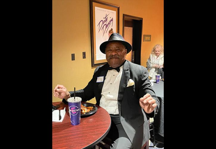 A smiling, well-dressed individual wears a black tuxedo and matching fedora.