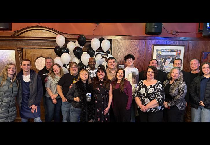 A large group takes a photo in a bar or restaurant setting. A person in the back holds up a party prop. Behind the group is a gathering of black and white balloons.