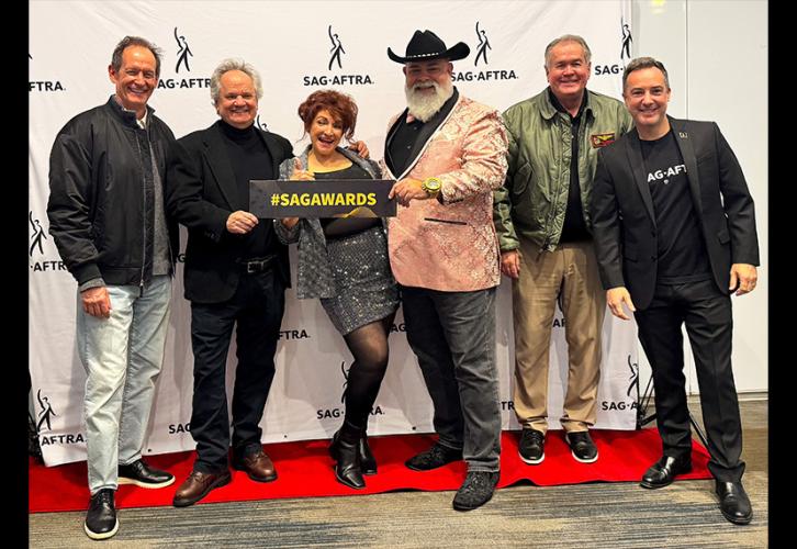 A small group of people stand on a red carpet in front of a white SAG-AFTRA step-and-repeat backdrop. Two people hold up a placard with “#SAGAWARDS” written in all-cap, yellow lettering.