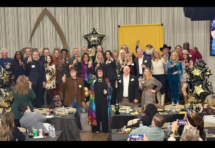 A wide shot of a very large group. Each person holds up a glass in a toast. Some other attendees are visible in the photo’s foreground. Also visible are some party decorations, largely balloons, and a yellow standing banner.