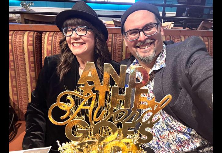 Two individuals are seated in a booth. The person on the left wears a black hat and black jacket; the person on the right wears a dark gray knit cap, colorful button-down shirt and grey jacket. In front of them is a gold centerpiece that reads, “And the award goes to” in gold. “Award” is in stylized text with flourishes and stars.