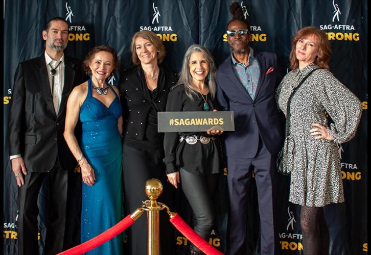 Six individuals are on the red carpet. In the foreground is a roped barrier; in the background is a black “SAG-AFTRA Strong” step-and-repeat backdrop. A person in the group holds a black placard with “#SAGAwards” in all-cap, yellow lettering.