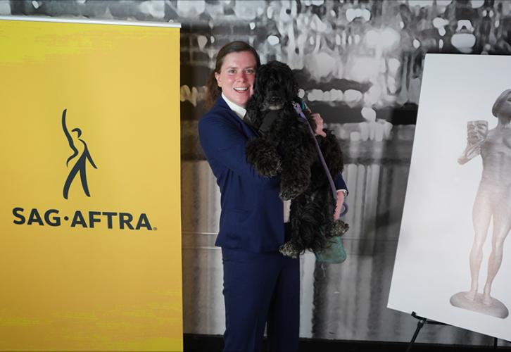 An individual in a blue suit holds a black dog. A yellow standing banner is on the left and a poster of The Actor statuette on an easel is on the right. 