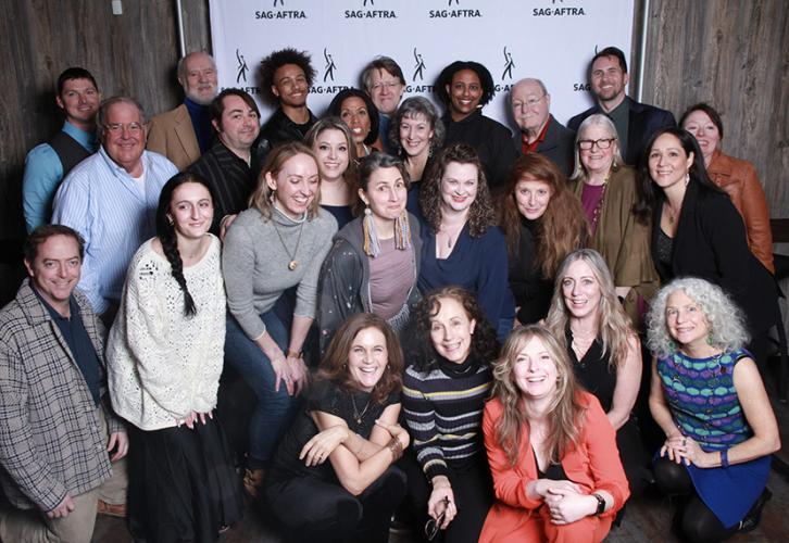 A large group of people gather for a photo inside of a nondescript room. Those in the first row are kneeling while the rest are standing. Behind the group is a white step-and-repeat backdrop with the SAG-AFTRA logo in black. 