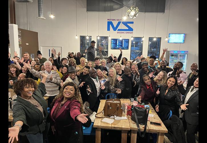 A large group of people gather inside an open office area. Each person makes the same pose with their arms open wide as if welcoming the viewer to join them.