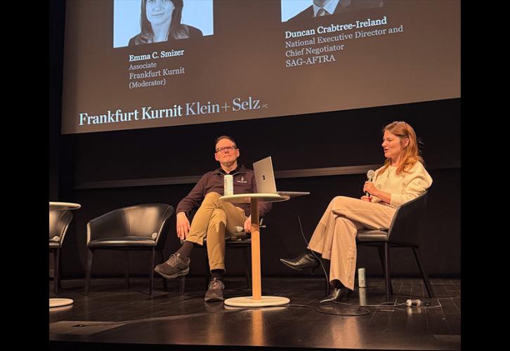 Crabtree-Ireland and Smizer are seated on stage. Between them on a small table is a laptop. Above them on a projector screen is a title card with their headshots and titles.