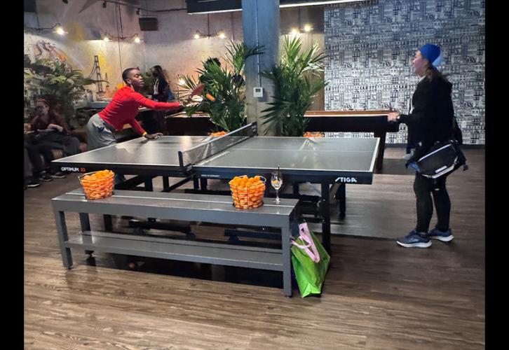 Two people play ping pong in a large recreational room. The person on the left is captured mid-play.