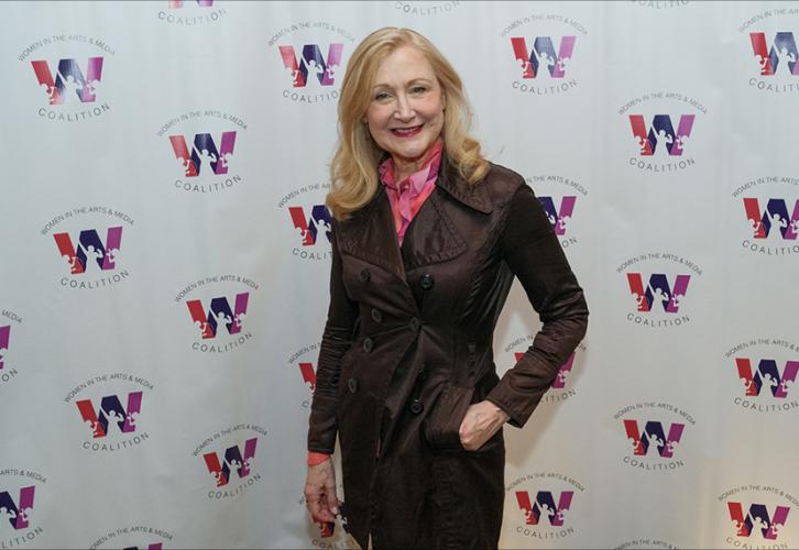Patricia Clarkson, smiling, is dressed in a dark jacket and pink blouse with their left hand in their pants pocket. Behind them is a white step-and-repeat backdrop with the Women in Arts & Media logo. 