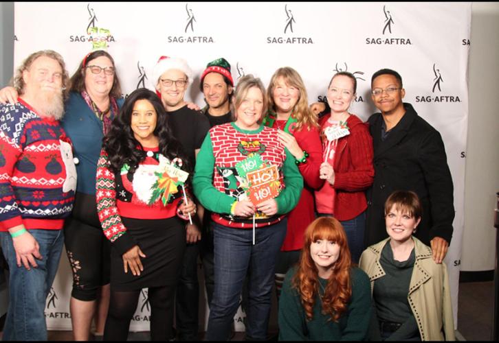 A group of individuals wearing holiday sweaters pose in front of a white backdrop with black SAG-AFTRA logos.