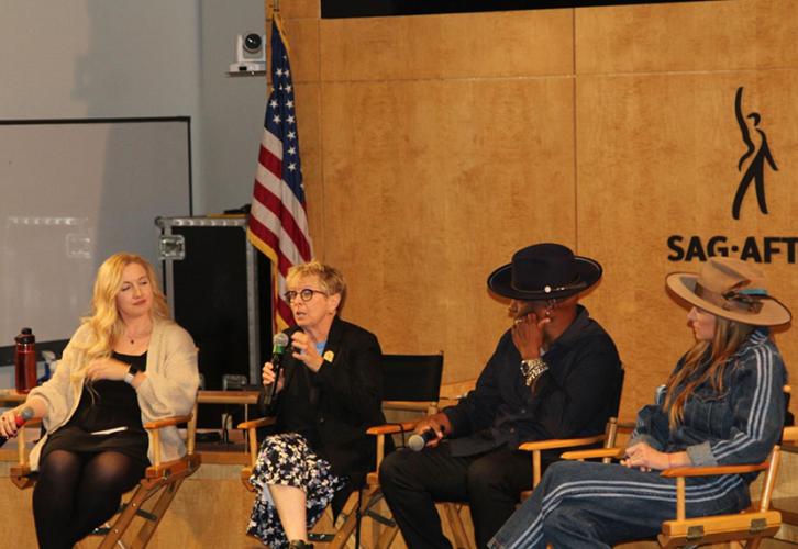Four individuals sit in directors chairs in front of a wood-paneled wall with the Sag-Aftra logo — a black figure with an upraised right arm with “Sag-Aftra” underneath. There is an American flag in the background.