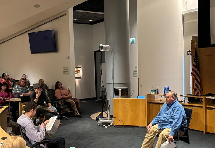 Two individuals sit in chairs acting out a scene with another individual sitting in a chair looking on, in front of an audience of people.