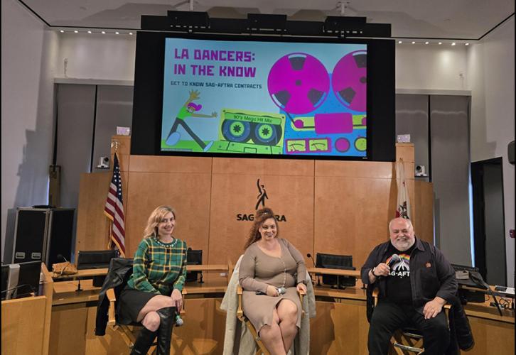 Three individuals sit in directors’ chairs with a colorful presentation behind them on a screen.
