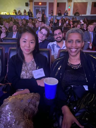 Eng, (Asian American), and Lyman (African American), are seated in theater chairs. Both women are dressed in party attire: Eng’s dress is gold; Lyman’s dress is black. There is a bright blue cup in the holder between them. In the background are others.