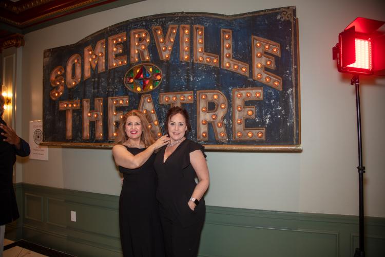 Aleman and Caruso stand closely together. Both wear all-black outfits. Aleman’s hand is on Caruso’s shoulder; Caruso’s hand is in her pocket. Behind them is a sign for the Somerville Theatre: light-up marquee letters are carved into blue marble.