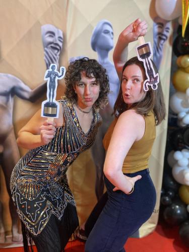 Xiadani and Curri stand close together. Xiadani’s dress is black with gold beading; Curri wears a gold top and blue pants. Their poses are more modelesque, with Xiadani bent forward at the hip, and Curri’s left hand on her hip and her right above head.