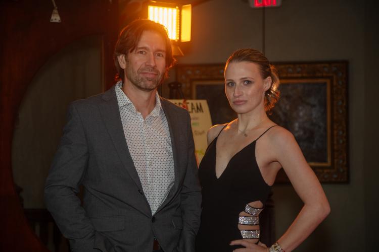 Gregorie and Renée stand together. Gregorie wears a gray blazer and white-patterned dress shirt; Renée wears a black dress with silver bands in the cutouts on the side. Renée’s left hand is on her hip. Behind Gregoire’s head is a warm gold light.
