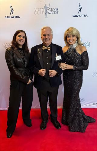 Madison, Garland and McElroy stand together on the red carpet. Madison and Garland wear black suits; McElroy’s dress is black with silver applique, and she wears earrings. McElroy’s arm is looped around Garland’s.