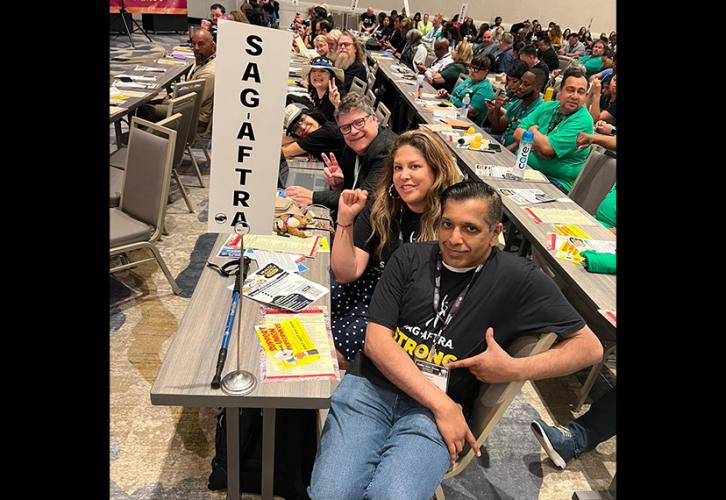 SAG-AFTRA members are seated at a table in a large conference room. In the foreground, two men and a woman look toward the camera. They each wear a black SAG-AFTRA-branded T-shirt. A stand holding a sign “SAG-AFTRA” has been placed at the table’s edge.