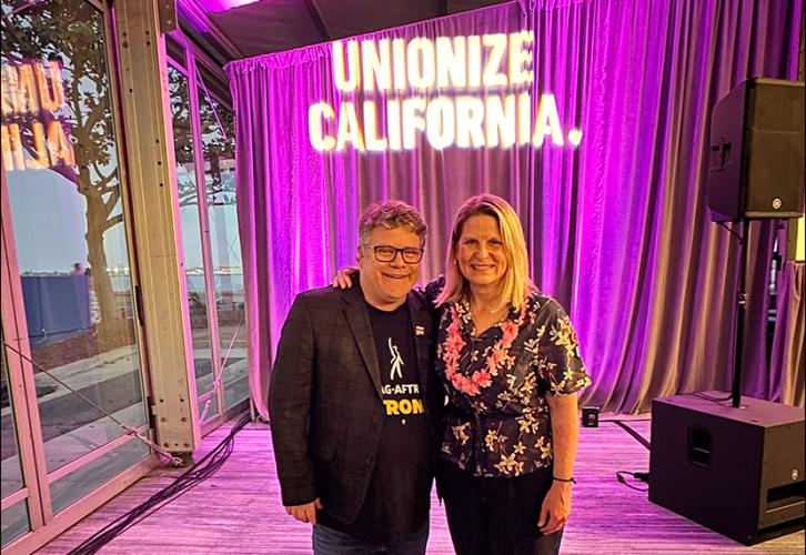 Astin wears a black blazer, black “SAG-AFTRA Strong” T-shirt and blue jeans; Shuler wears a flower-patterned shirt and Hawaiian lei with dark slacks. Shuler’s arm is around Astin’s shoulders. Behind them is a projection saying “Unionize California."